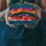 person holding meat sandwich with sliced tomatoes, onions, and grilled meat