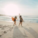 two women walking towards the ocean carrying surfboards during day - lesbian holidays