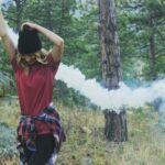 woman raising arms holding white smoke bomb standing beside rock surrounded with trees at daytime