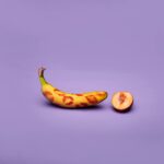 A whimsical arrangement featuring a banana with kiss marks and a peach half on a purple surface.