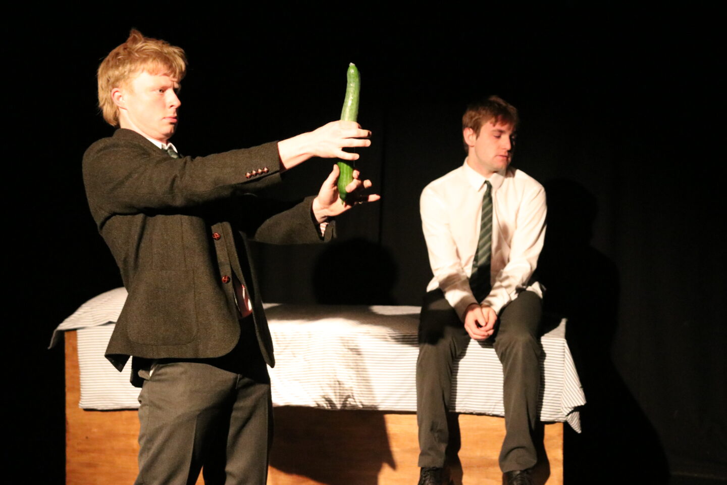 a man holding a cucumber and another man sits on a bed