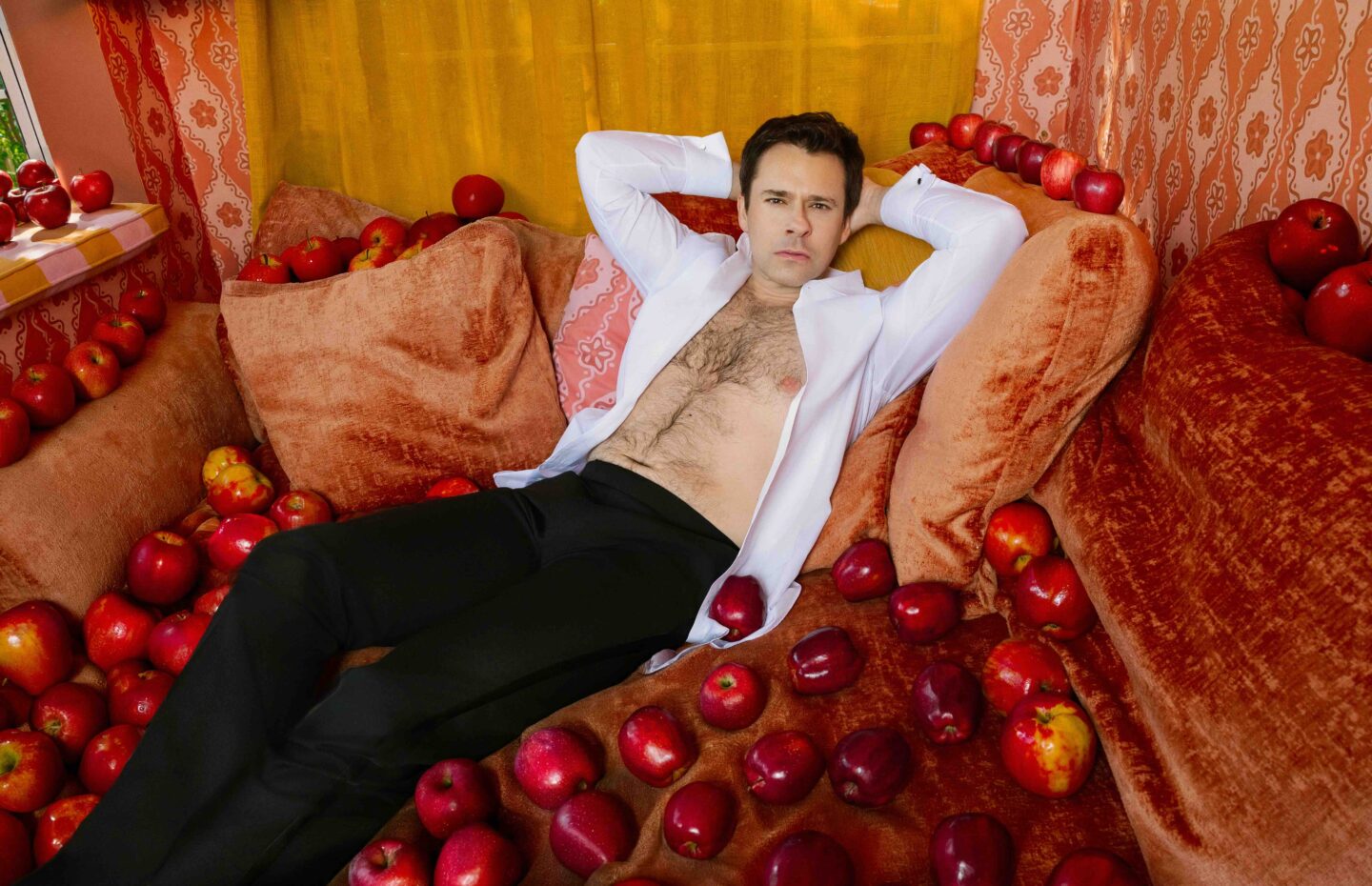 a man with his shirt open, laying on a couch surrounded by apples