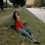 Two women share a tender moment by a riverside, surrounded by nature and peaceful scenery.