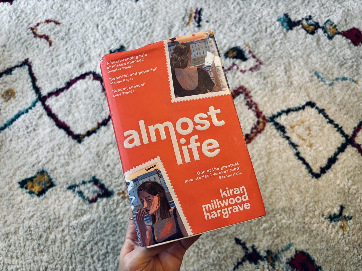 A hand holding a book, Almost Life, with a pinky orange cover and two illustrations of girls on the front cover.